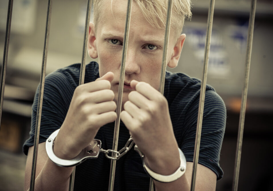 Rebellious teenager taken prisoner by the police standing glaring at the camera from behind bars with handcuffed hands